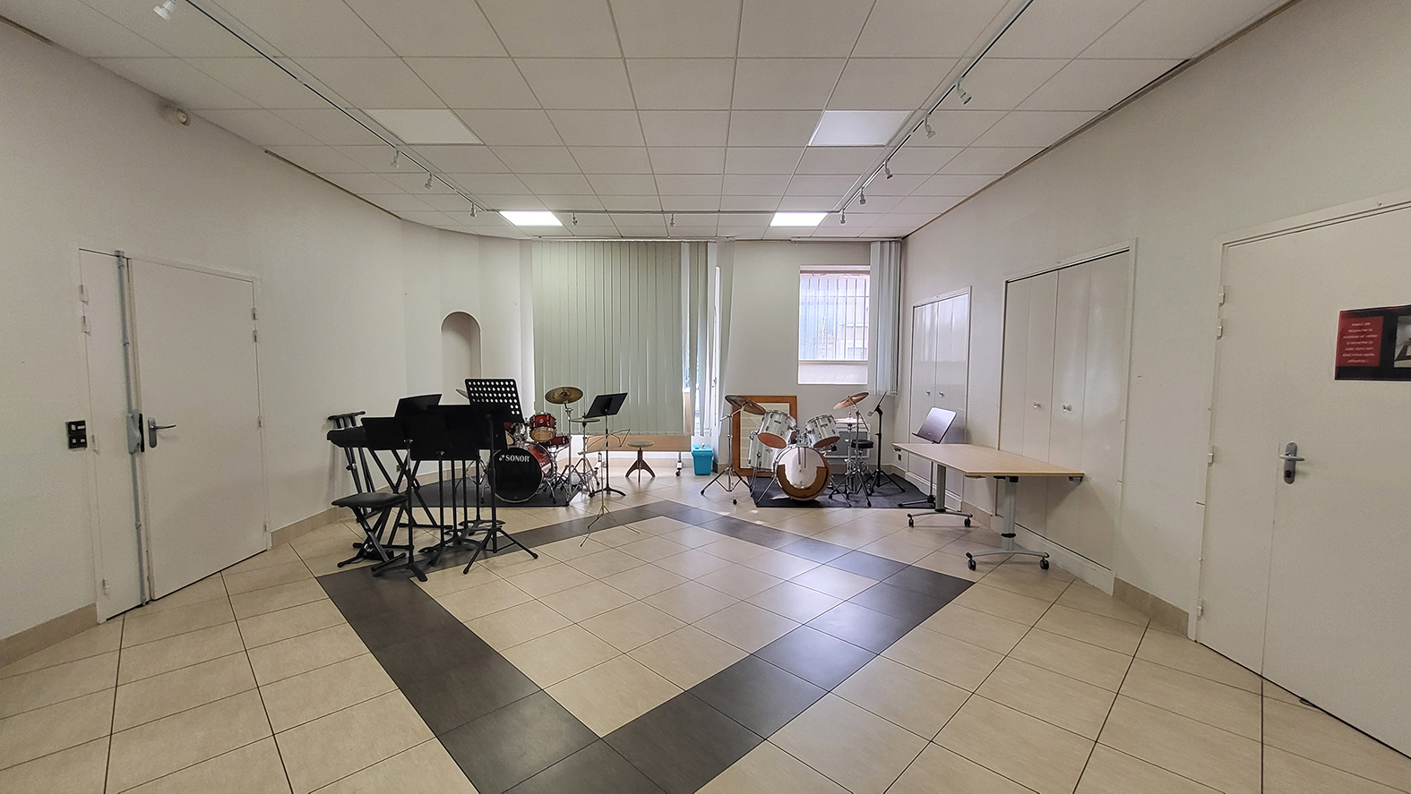 Salle avec batterie, piano, pupitres, tables et chaises.