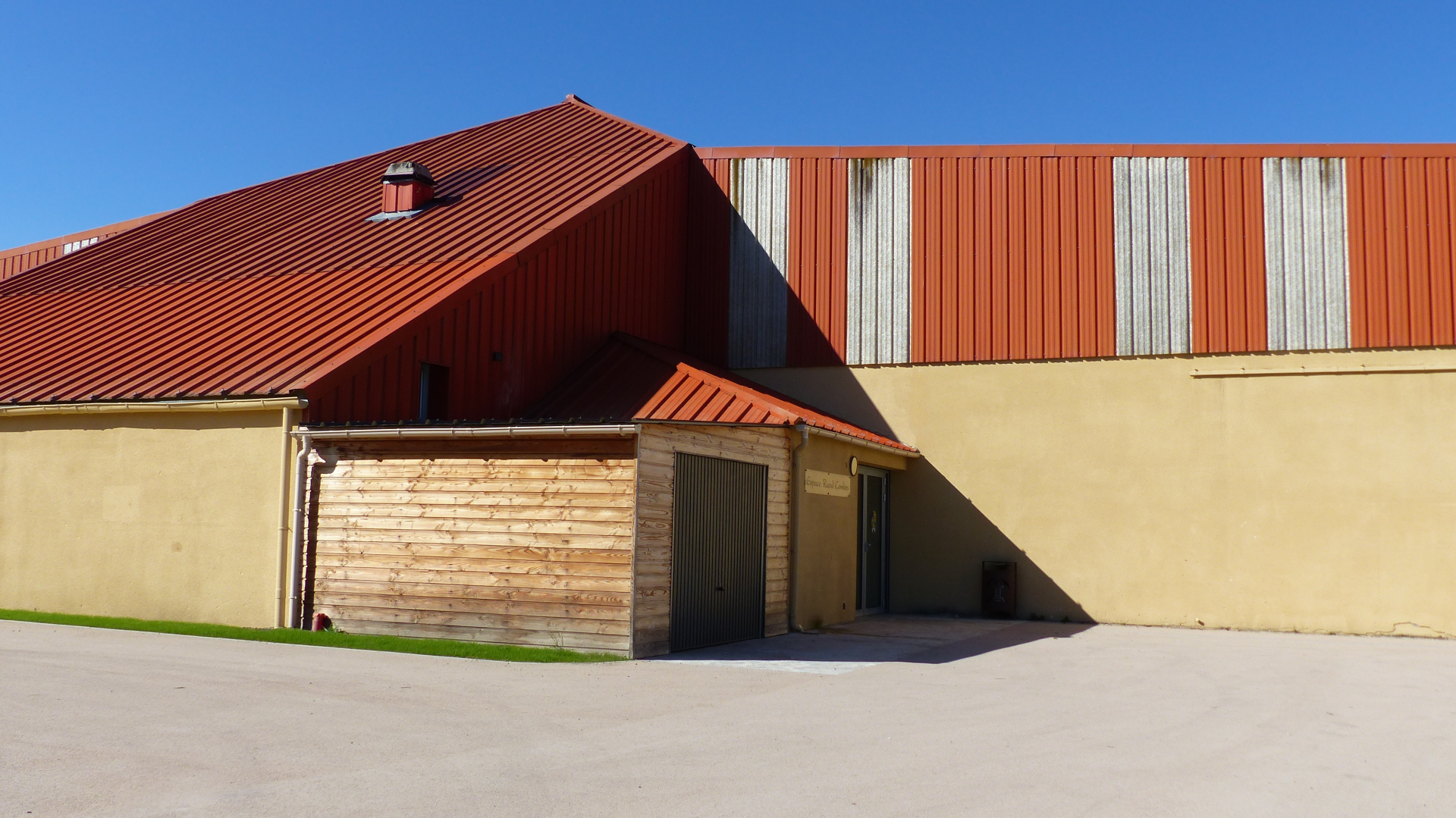 Bâtiment industriel avec toit rouge, murs beiges et orange, porte de garage et cheminée.