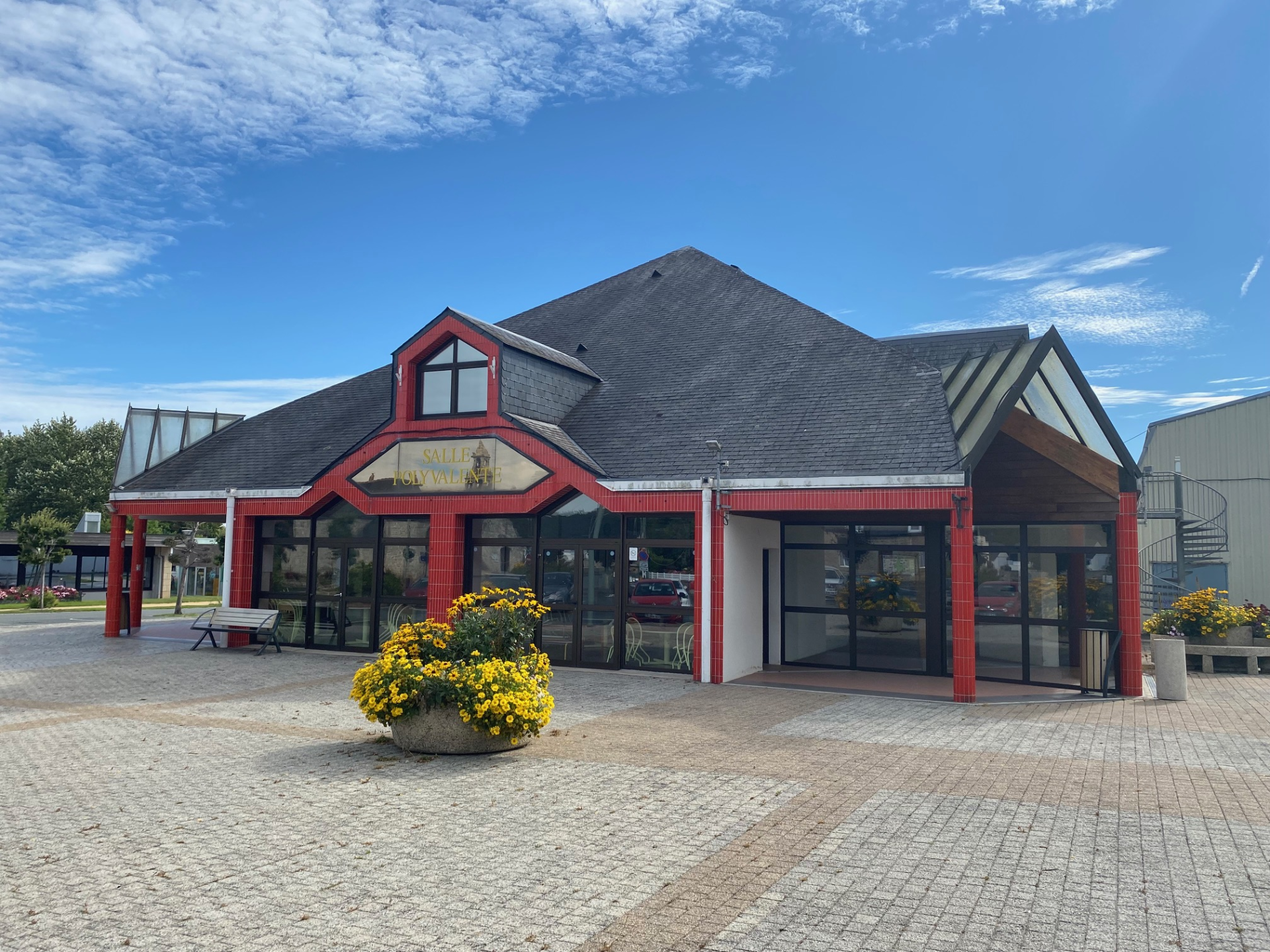 "Bâtiment avec pignons, grandes fenêtres, porte d'entrée, bancs, pots de fleurs, et ciel dégagé."
