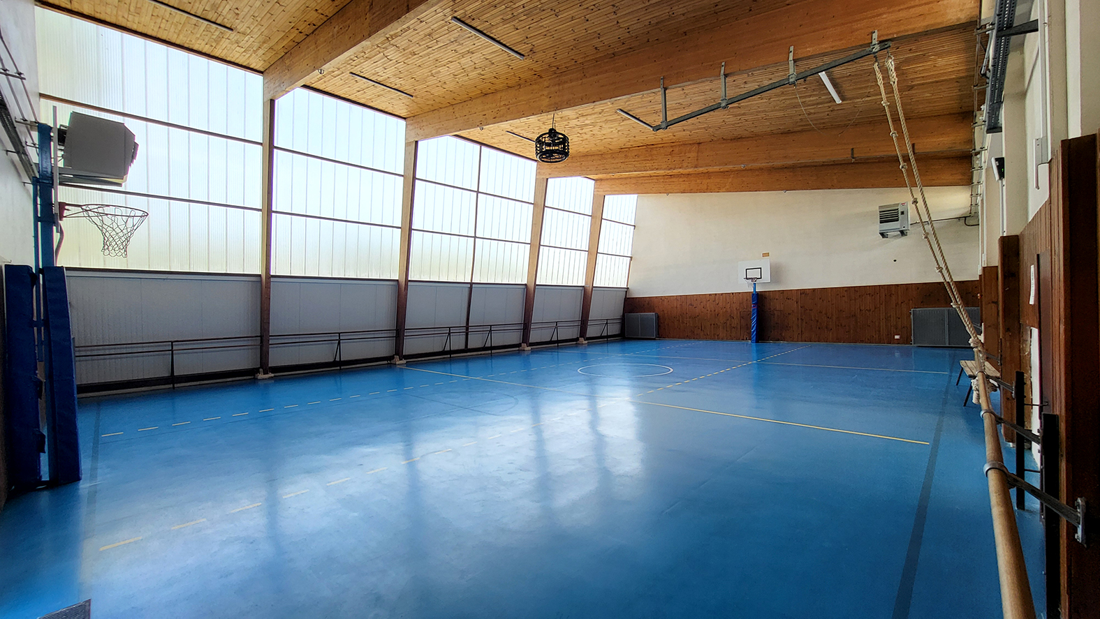 Salle de sport avec paniers de basket, murs en bois, parquet bleu et grandes fenêtres.