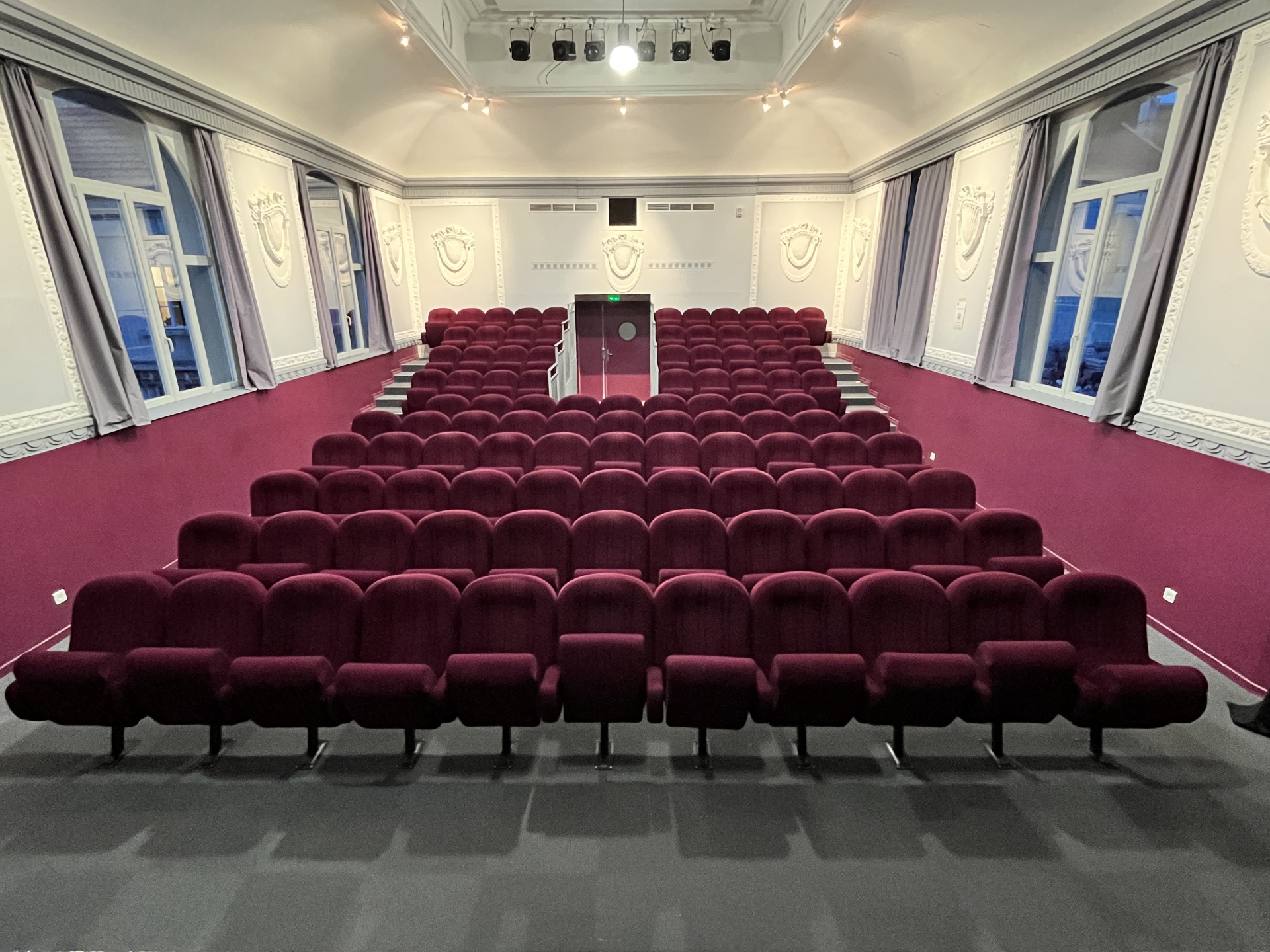 Salle avec rangées de sièges rouges, écran, projecteur et lumières au plafond.