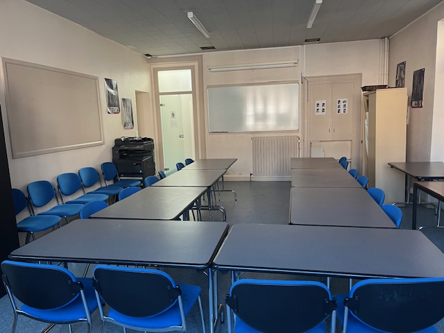 Salle de classe avec tables, chaises bleues, tableau blanc, porte, radiateur et tableau d'affichage.