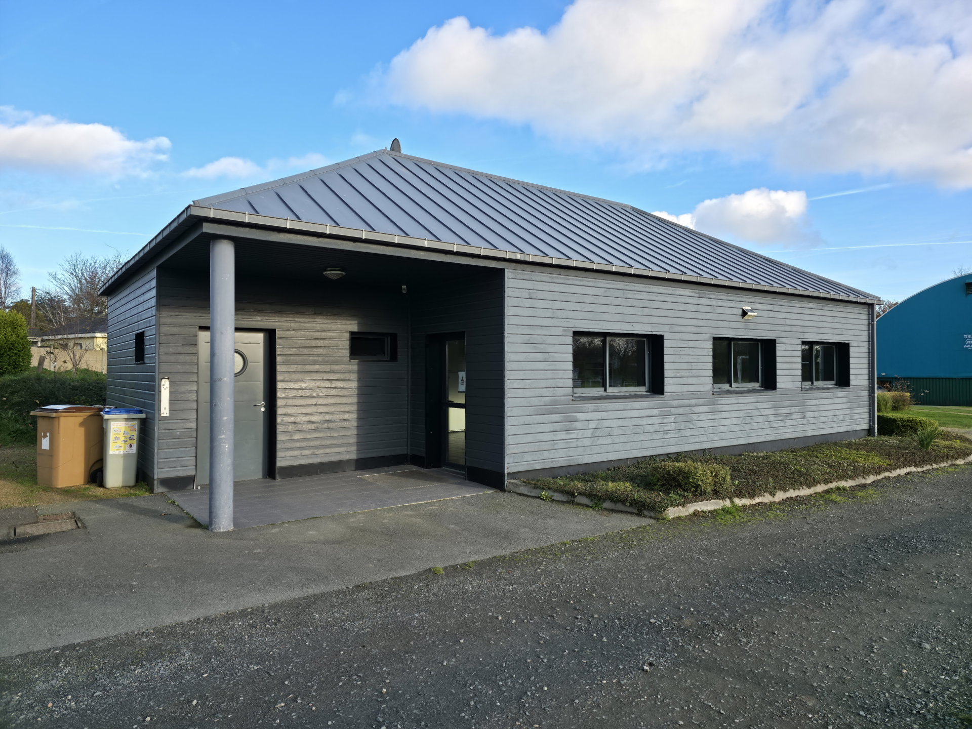 Bâtiment gris avec toit plat et colonnes, portes et fenêtres. Poubelles à côté. Ciel bleu avec nuages.