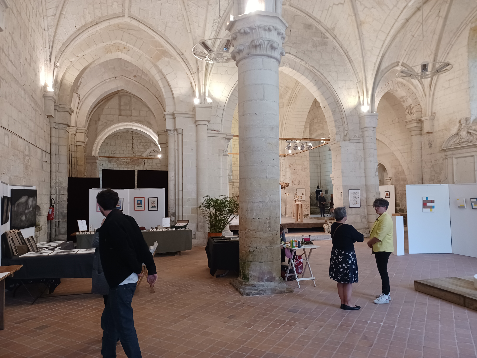 Salle voûtée avec colonnes, tables, chaises, présentoirs, œuvres d'art, et personnes.
