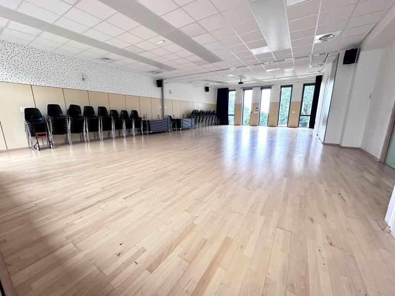 Salle avec parquet, chaises alignées, murs décorés, portes vitrées.