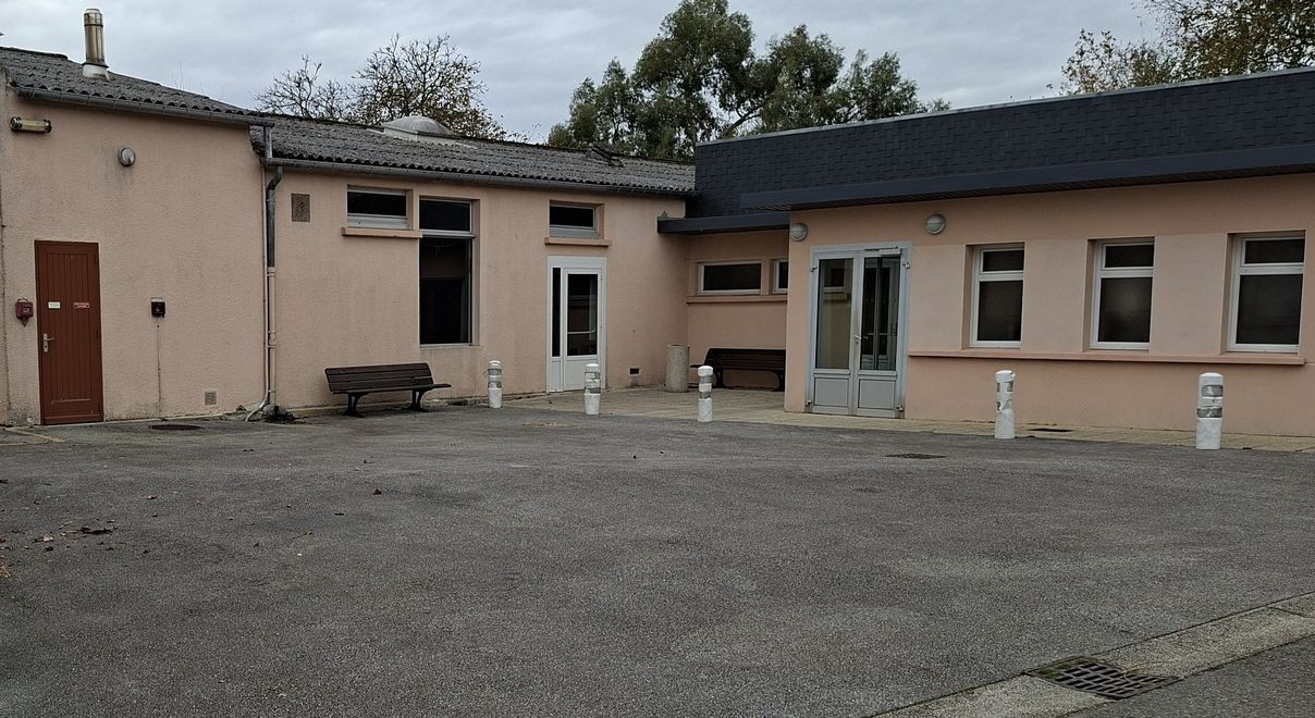 Bâtiment beige avec plusieurs fenêtres, porte en bois, banc et barrières.