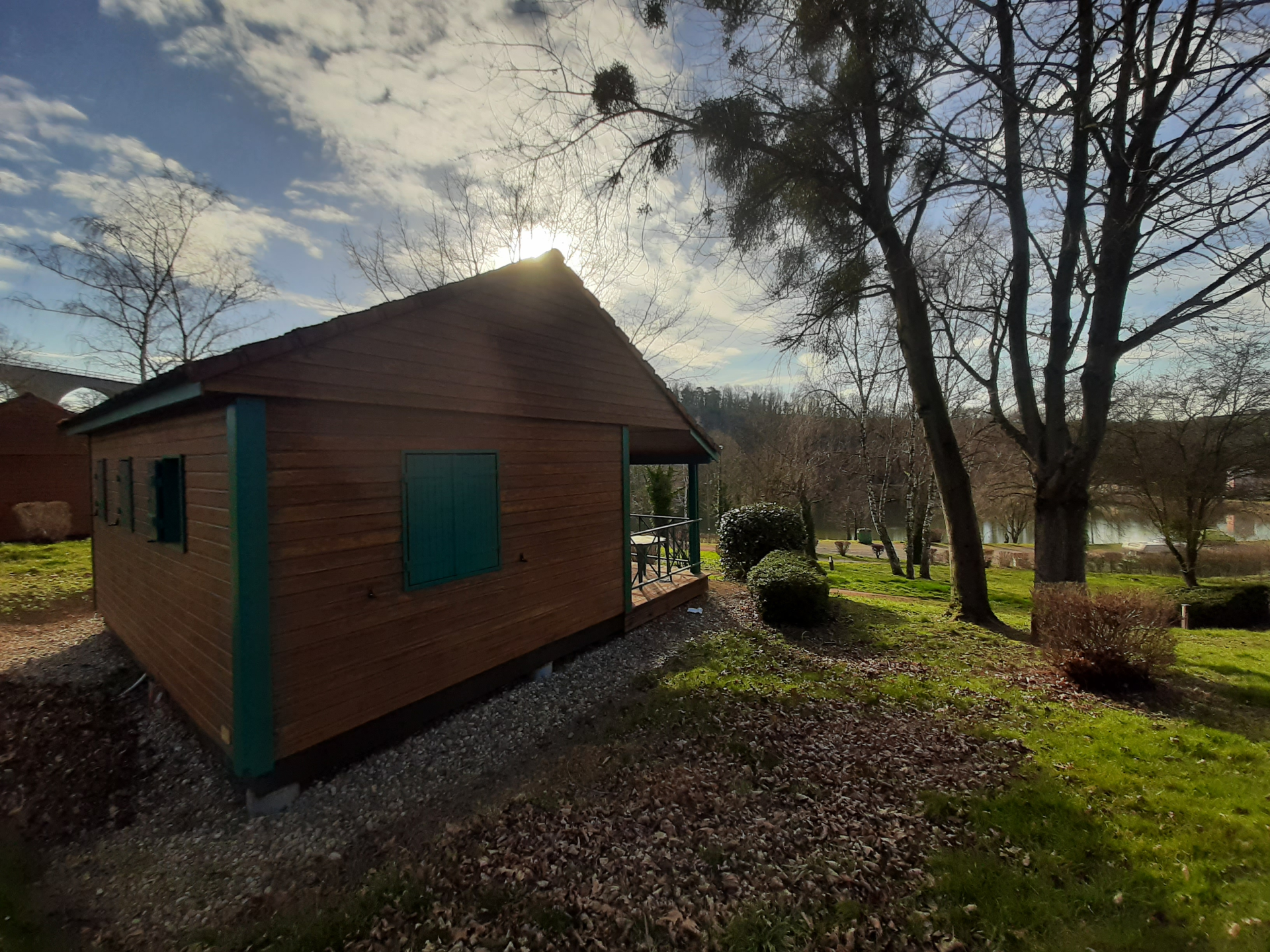 Cabane en bois avec fenêtres vertes, située dans un jardin avec arbres et gravier au sol.