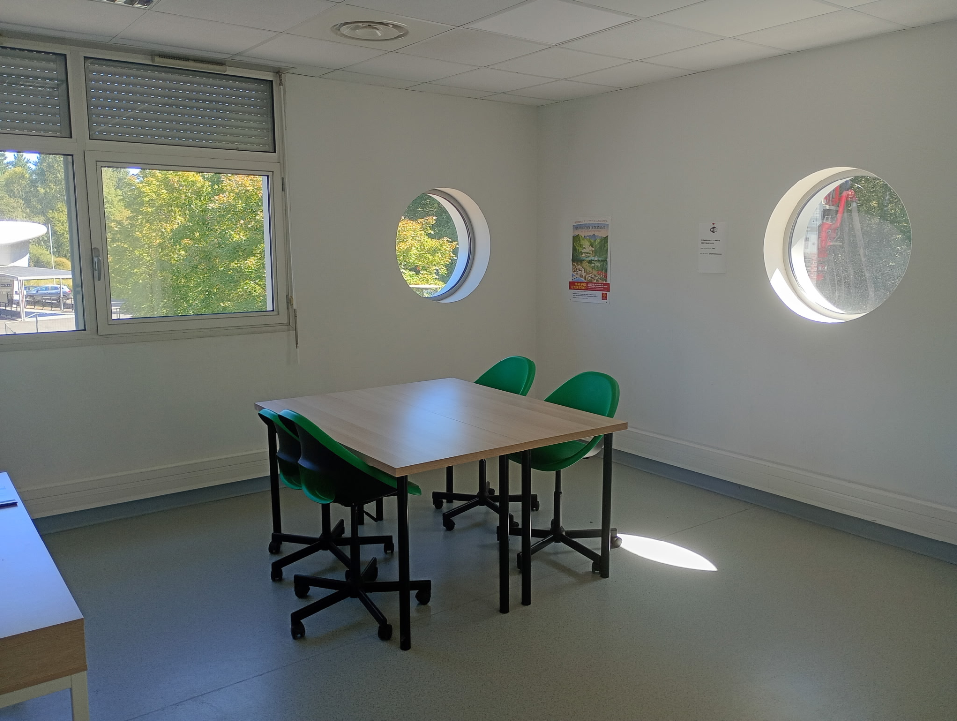 Salle avec table, chaises, fenêtres rondes et rectangulaire, affiches.