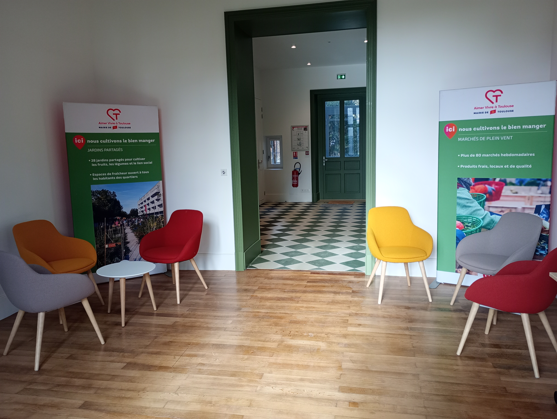 Salle d'attente avec chaises colorées, tables basses, panneaux d'affichage et porte verte.