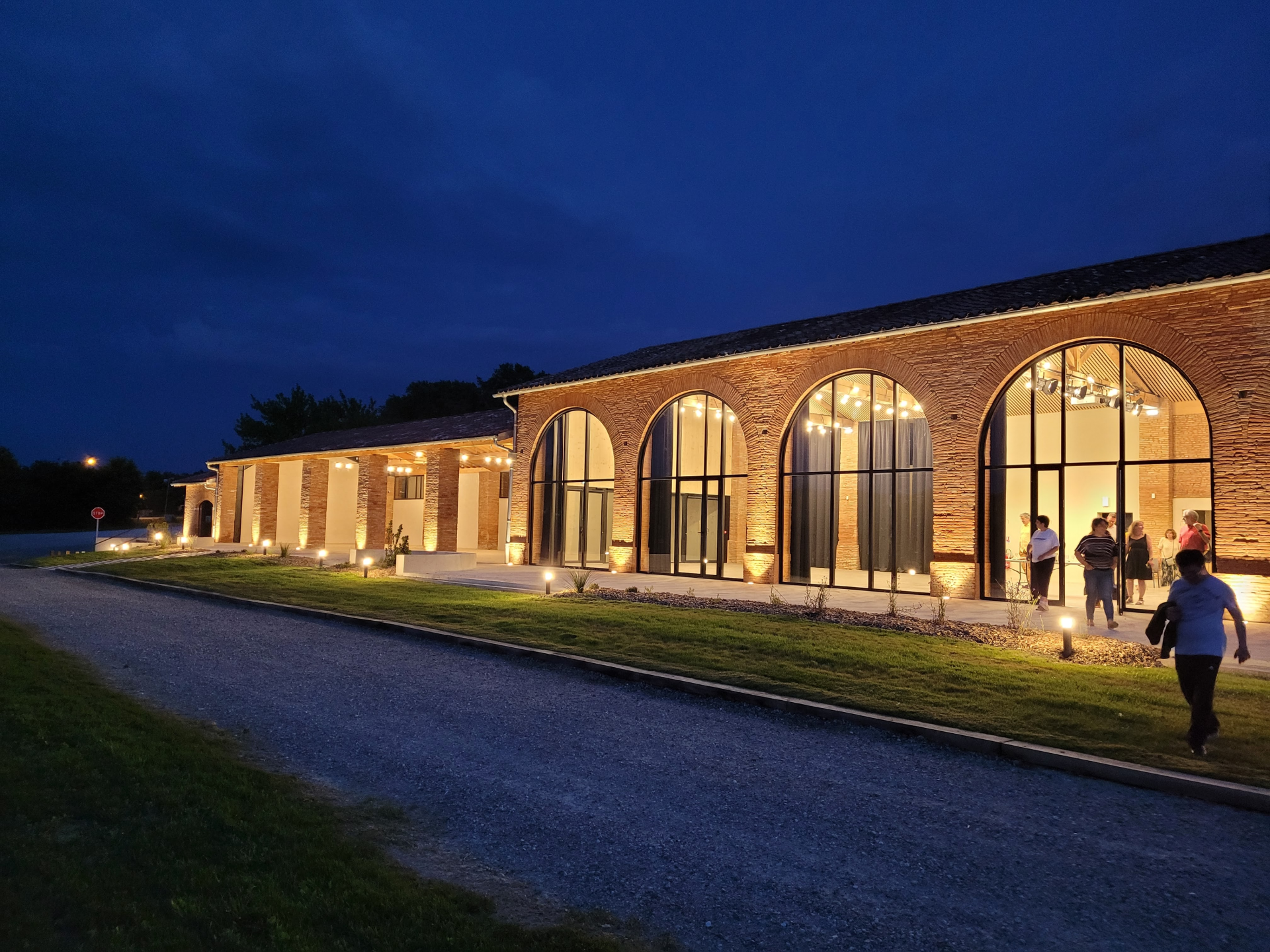 Bâtiment en brique avec grandes fenêtres éclairées, allée et pelouse.