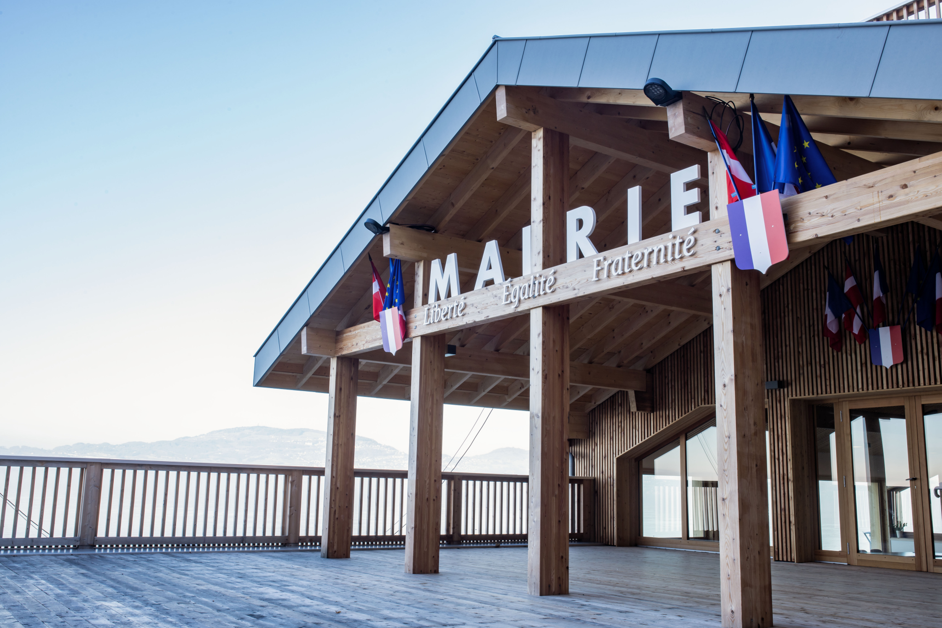 Bâtiment en bois avec enseigne "Mairie Liberté Egalité Fraternité", drapeaux, balcon et vue sur montagnes.