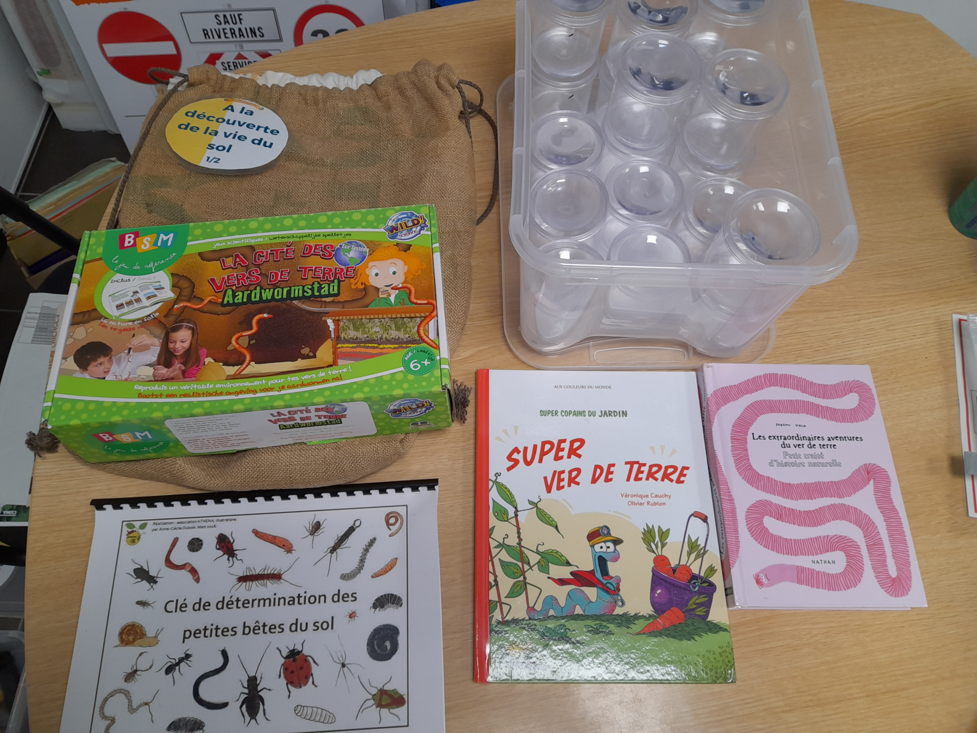 Sur une table, un sac en toile, une boîte de 24 pots transparents, un livre, un dépliant, une clé de détermination des petites bêtes du sol et un stylo.