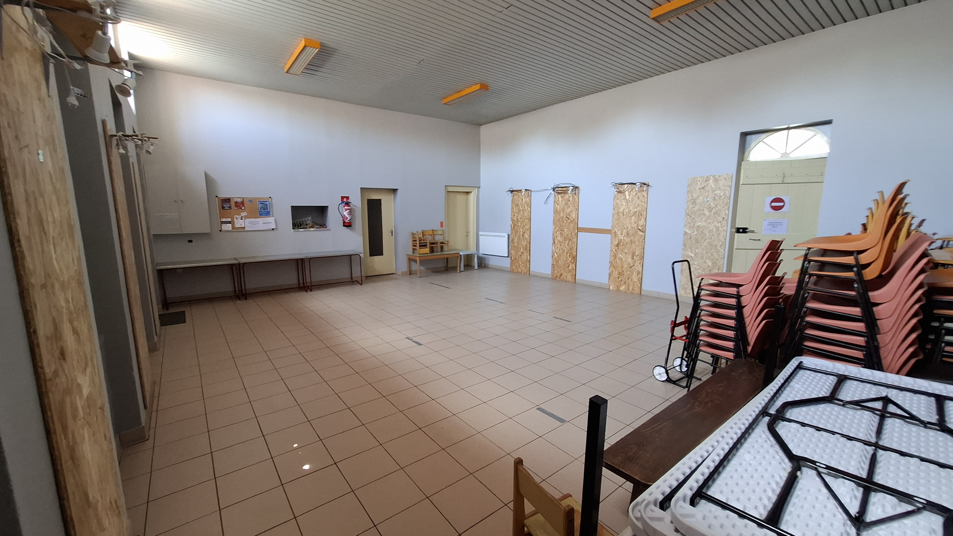 Salle avec tables, chaises empilées, chariot, et matériel de rangement.