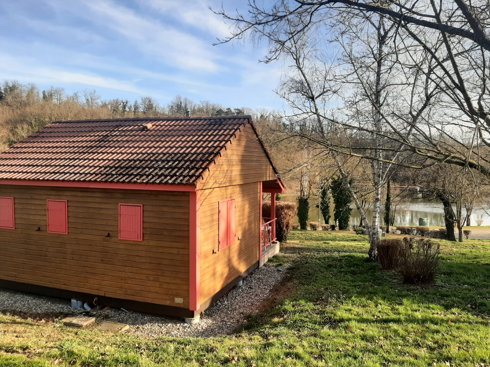 Petite maison en bois avec toiture en tuiles, portes rouges, située dans un jardin avec arbres et autre habitation au loin.