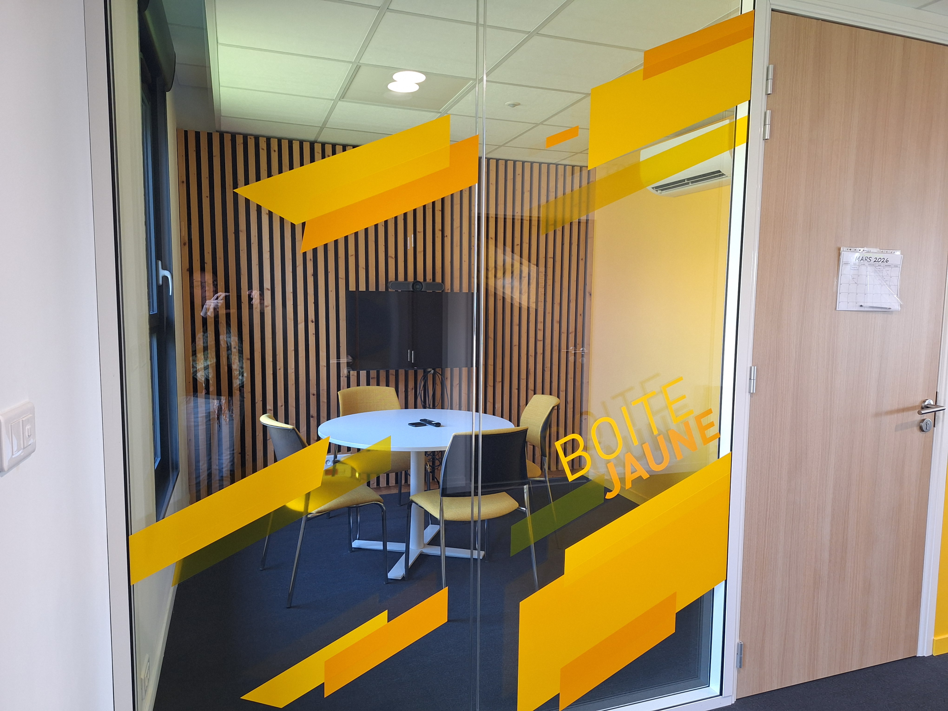 Salle de réunion avec table ronde, chaises, écran. Porte en verre avec motifs jaunes.