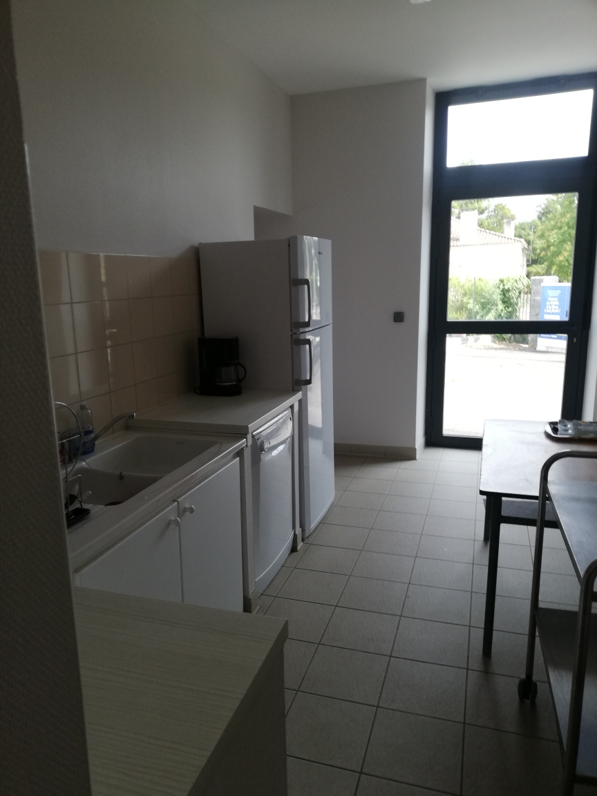 Cuisine avec évier, réfrigérateur, four, micro-ondes, table et chaises. Porte vitrée menant à l'extérieur. Carrelage au sol.