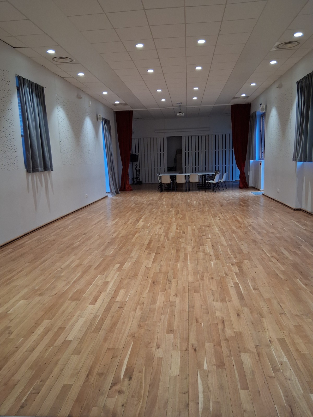 Salle vide avec parquet, tables, chaises et rideaux.