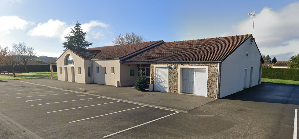 Bâtiment avec toiture en tuiles, façade mixte pierre et enduit, porte de garage blanche, parking marqué, arbres.