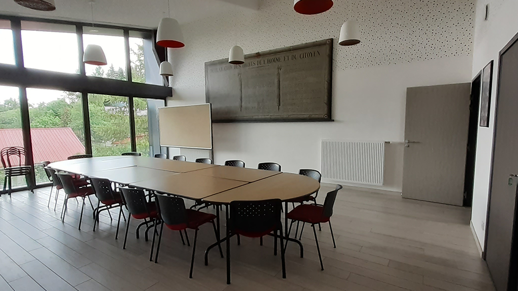 Salle avec table, chaises, tableau blanc, radiateur, porte et suspensions.
