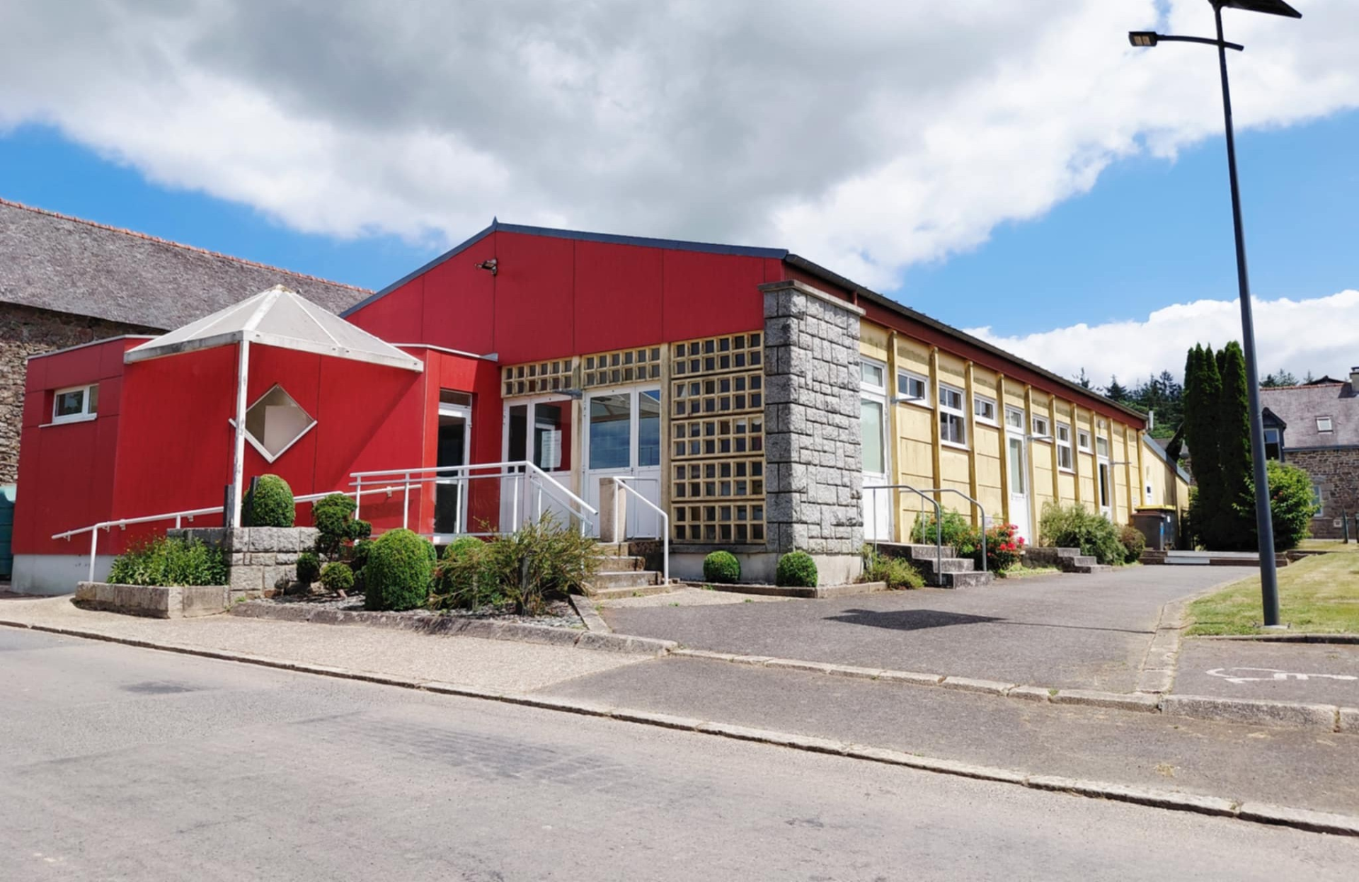 Bâtiment avec façade rouge et jaune, toiture grise, rampe d'accès, plantes et réverbère.
