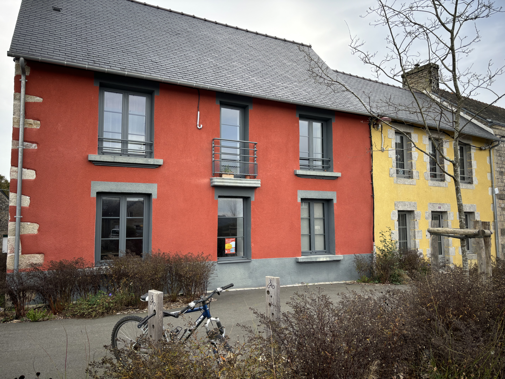 Maison rouge à deux étages avec balcons, vélo devant, arbustes et toits en ardoise.