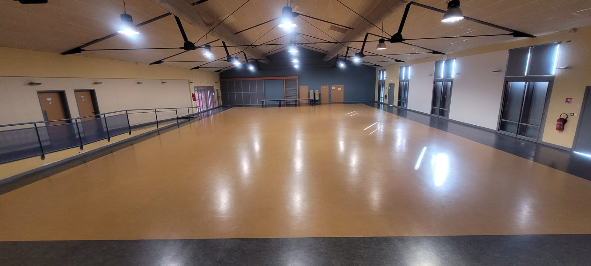 Salle spacieuse avec éclairage suspendu, sol en bois, mezzanine, portes et fenêtres.
