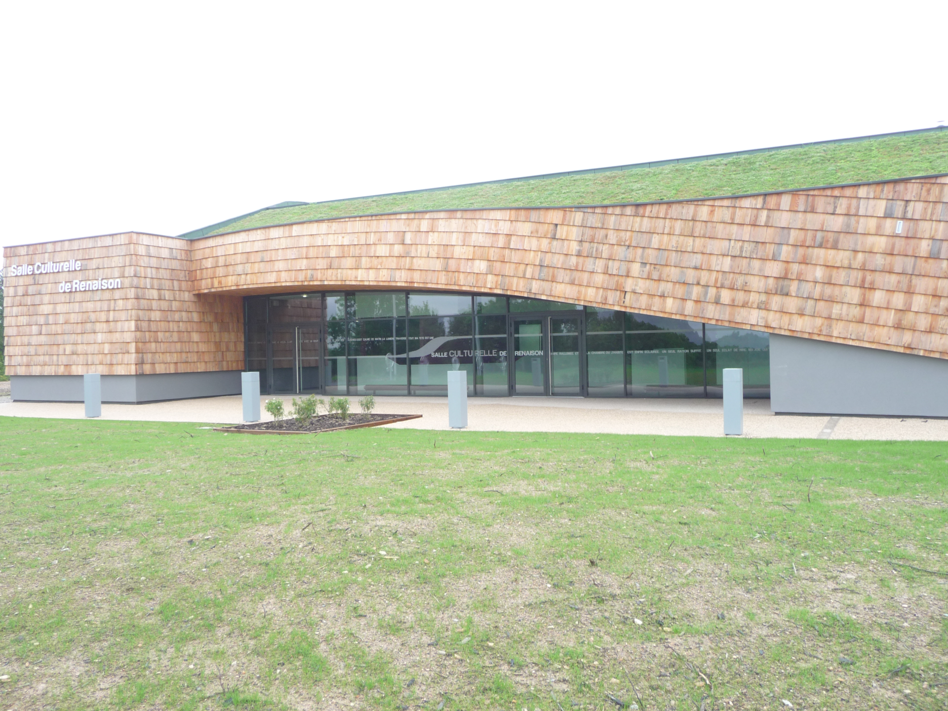 Bâtiment moderne avec façade en bois, toit vert, grandes vitres et pelouse.