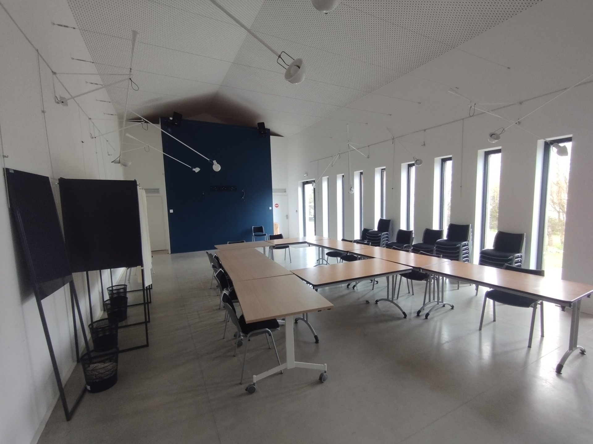 "Salle de réunion avec tables, chaises, tableau blanc, pot de fleurs, fenêtres, éclairage suspendu et porte."