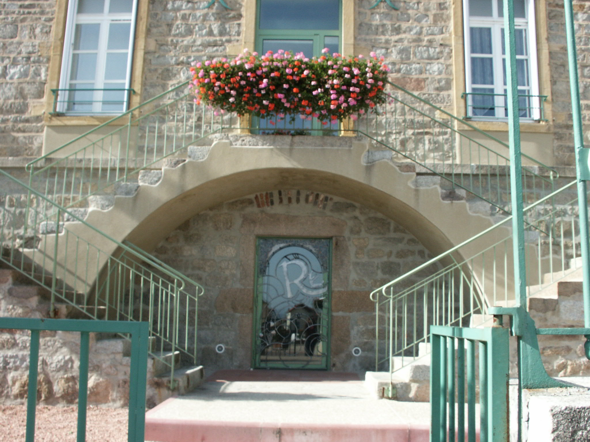 Entrée avec escalier, rambarde verte, porte en arc, vitrail et fleurs.