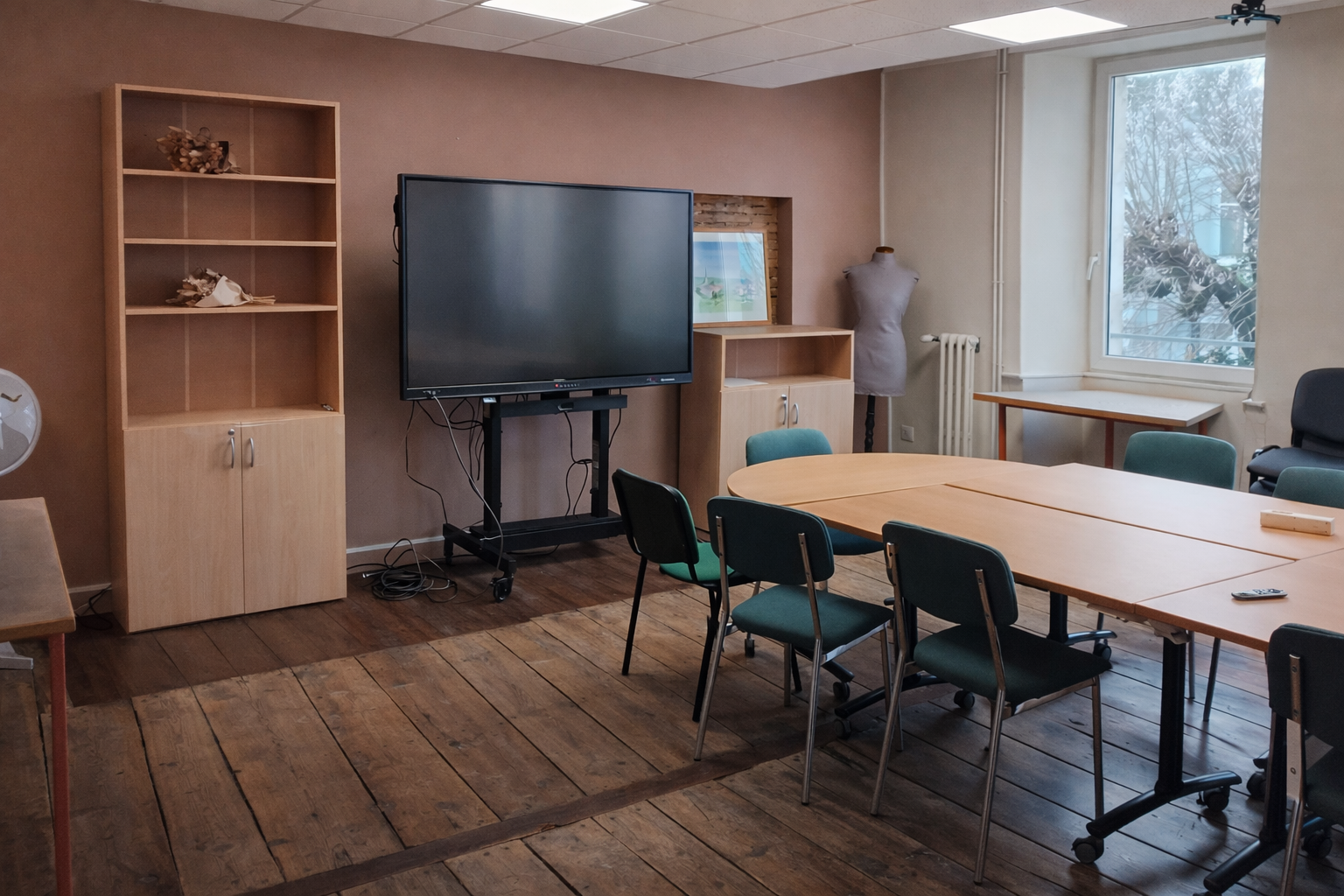 Salle avec écran, table, chaises, étagère, mannequin et radiateur.
