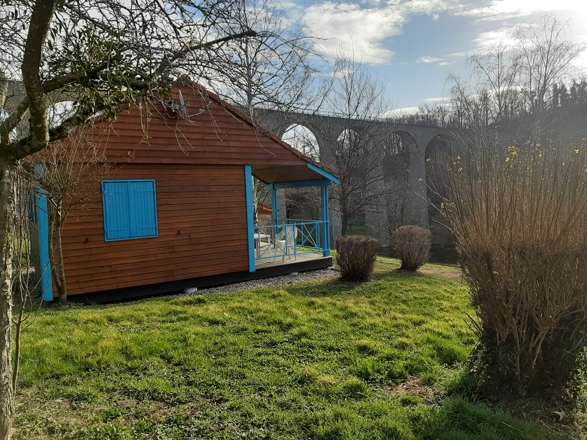 Pavillon en bois avec fenêtres bleues et terrasse, pont en pierre en arrière-plan, jardin avec herbe et buissons.
