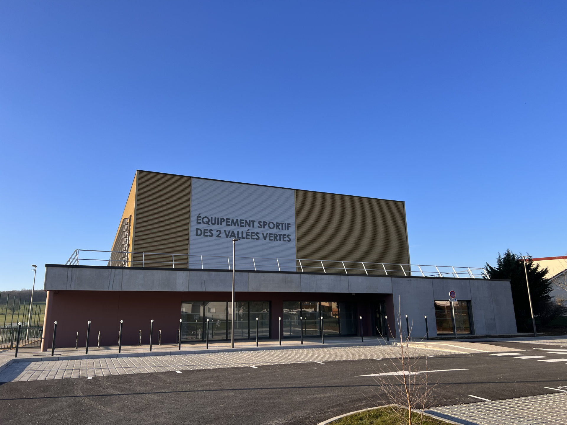 Bâtiment avec façade en bois et verre, parking et panneau "Équipement sportif des 2 vallées vertes".