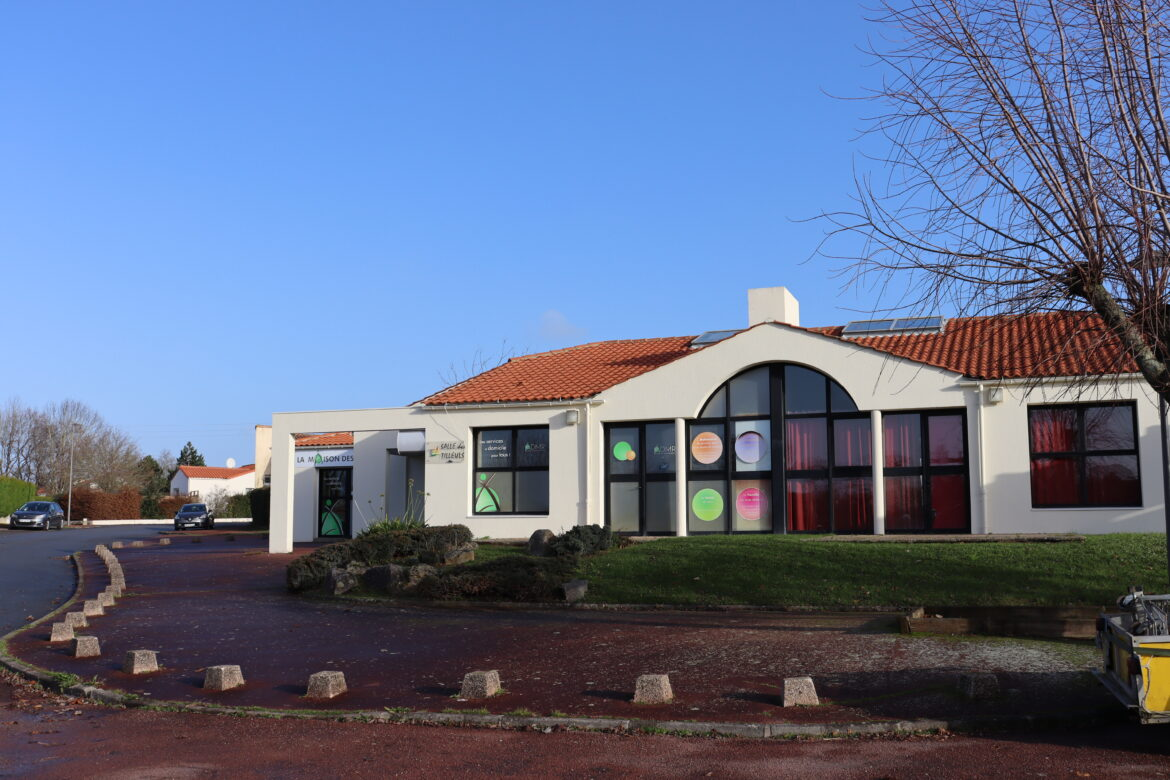 Bâtiment blanc avec toiture rouge, grandes fenêtres, parking et végétation.
