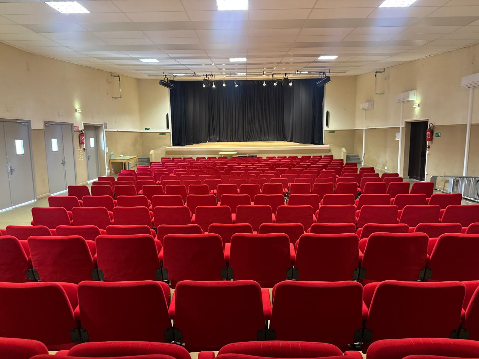 Salle avec rangées de sièges rouges, scène, rideau, éclairage et portes.