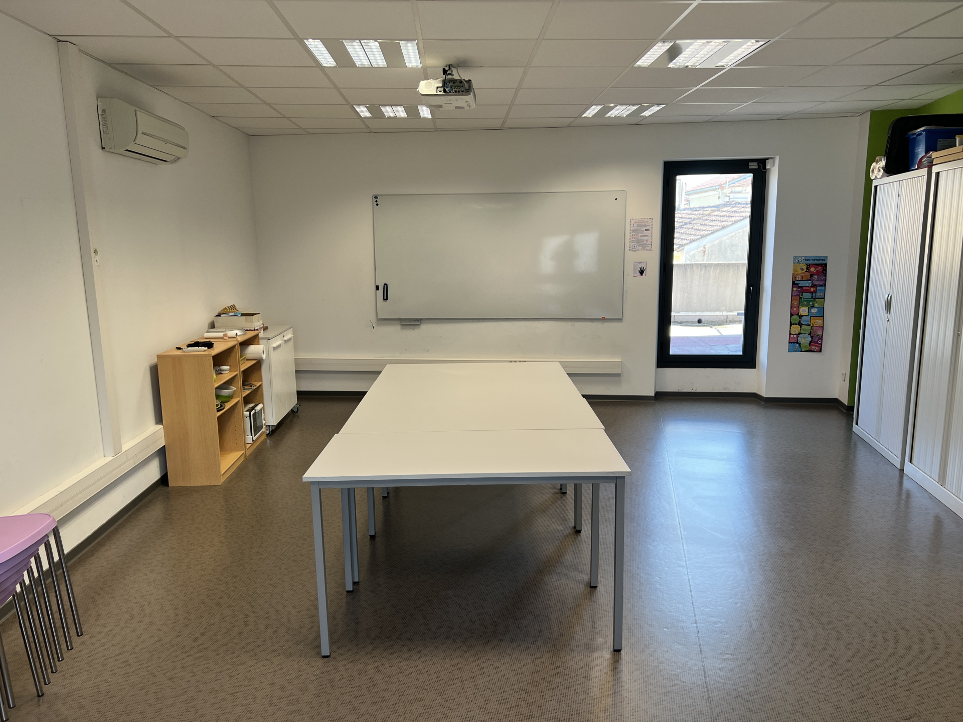 Salle avec table, chaises, tableau, étagères, vidéoprojecteur et climatiseur.