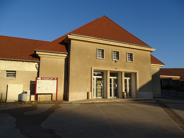 Bâtiment avec toit rouge, entrée principale en verre, un panneau de stationnement et une poubelle visible.