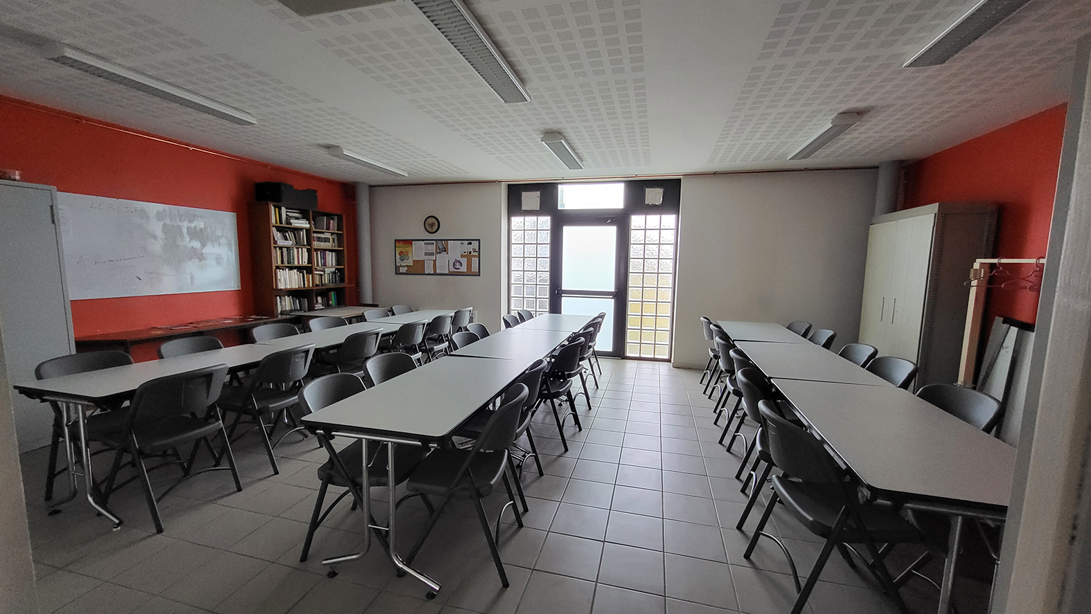 Salle avec tables, chaises, tableau blanc, étagères, porte et fenêtres.