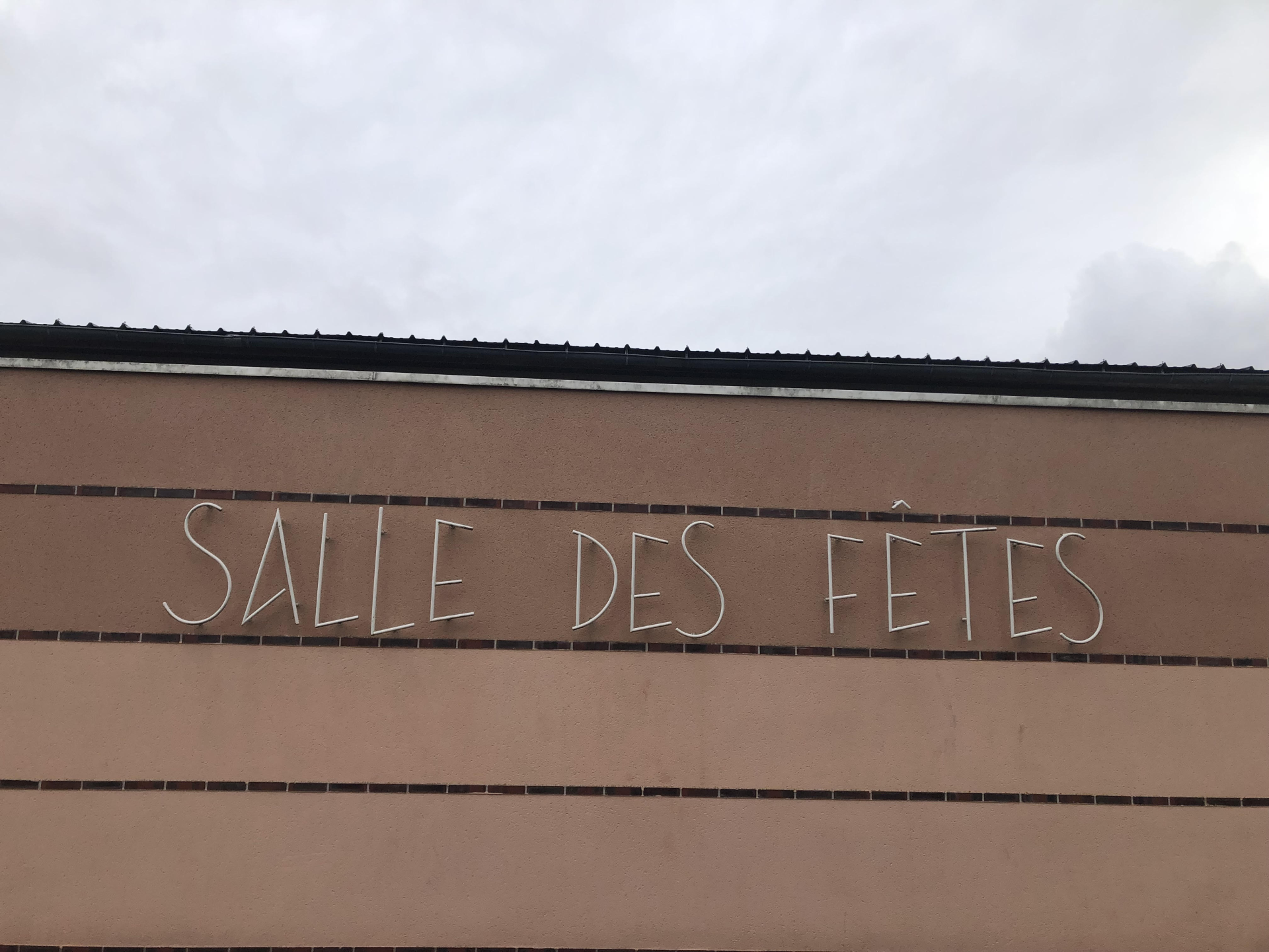 L'image montre une partie d'un bâtiment en béton avec l'enseigne "Salle des fêtes" accrochée au mur.