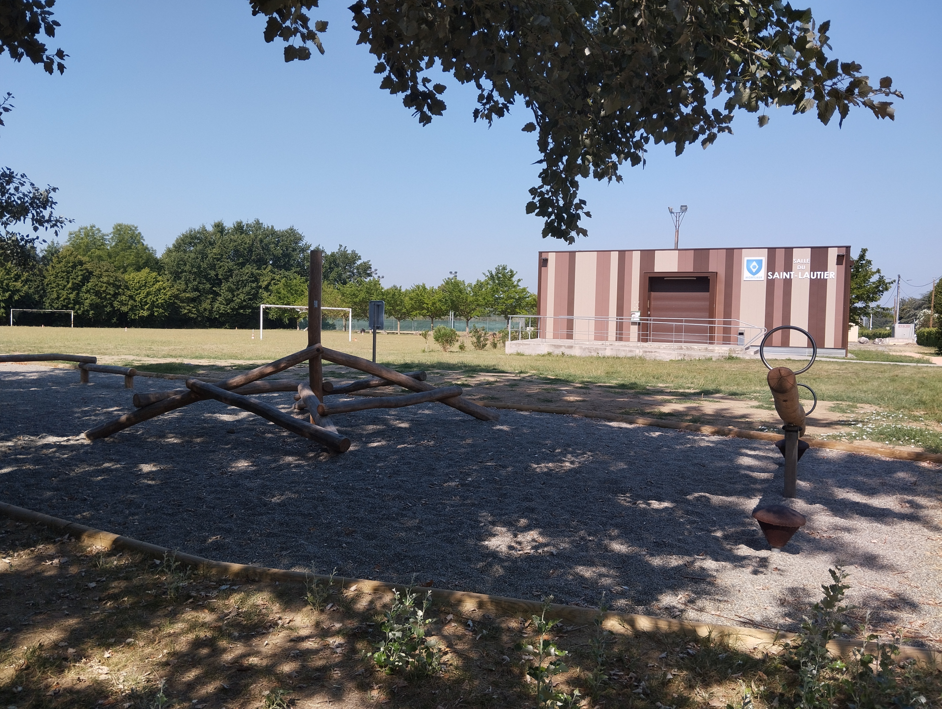 Terrain de sport avec panier de basket, structure en bois et bâtiment en arrière-plan.
