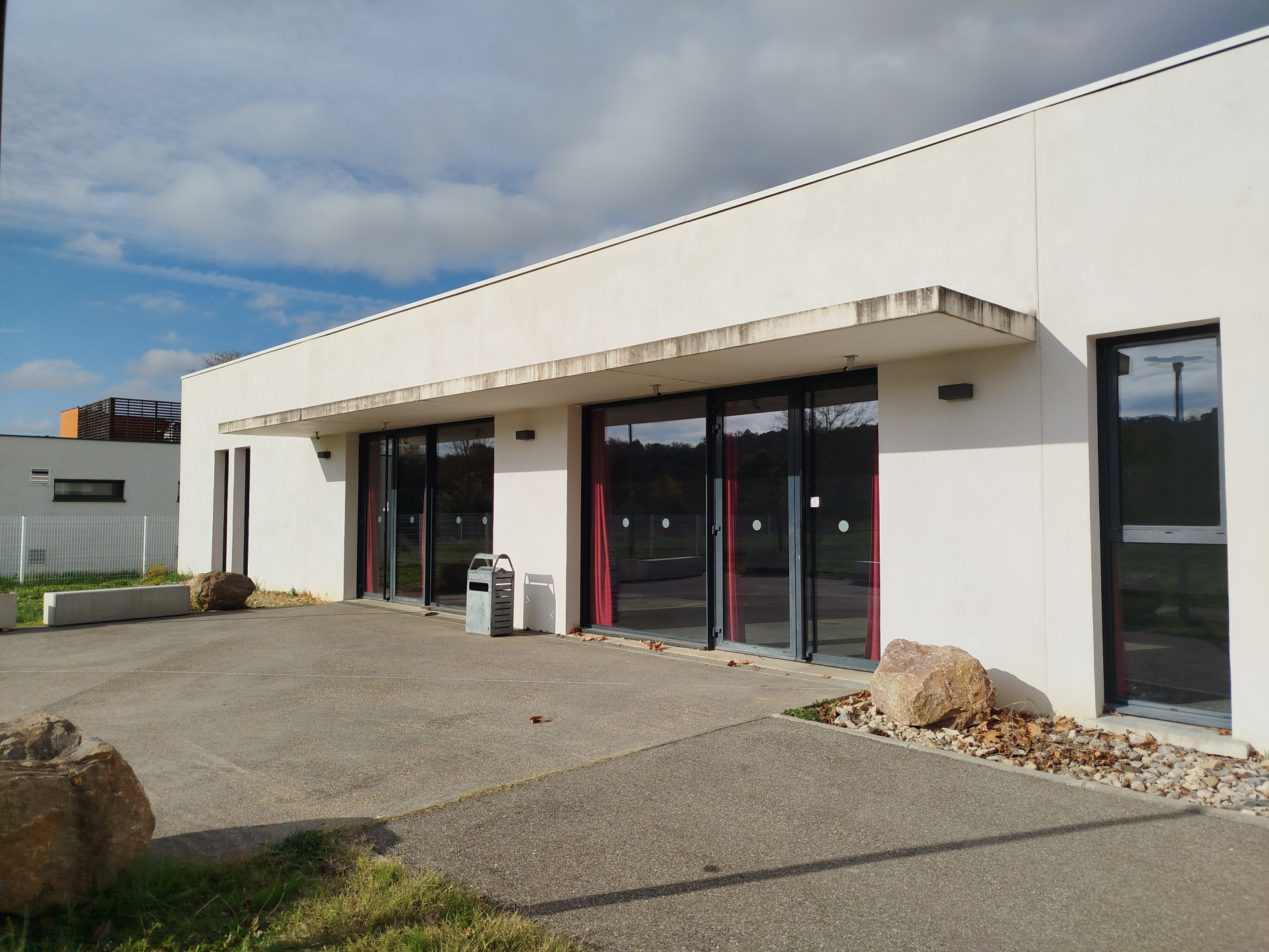 Bâtiment blanc avec grandes baies vitrées et portes, parking et rochers.