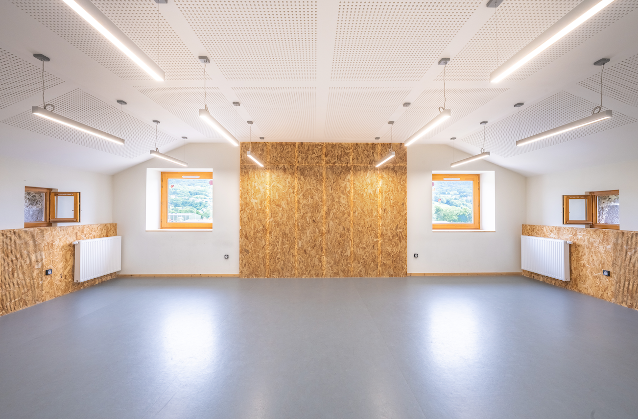 "Salle vide, plancher gris, murs partiel en bois, fenêtres, radiateurs, éclairage suspendu."