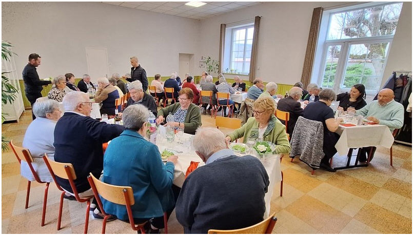 Salle avec tables, chaises, serviettes, verres et plantes. Personnes assises et debout.