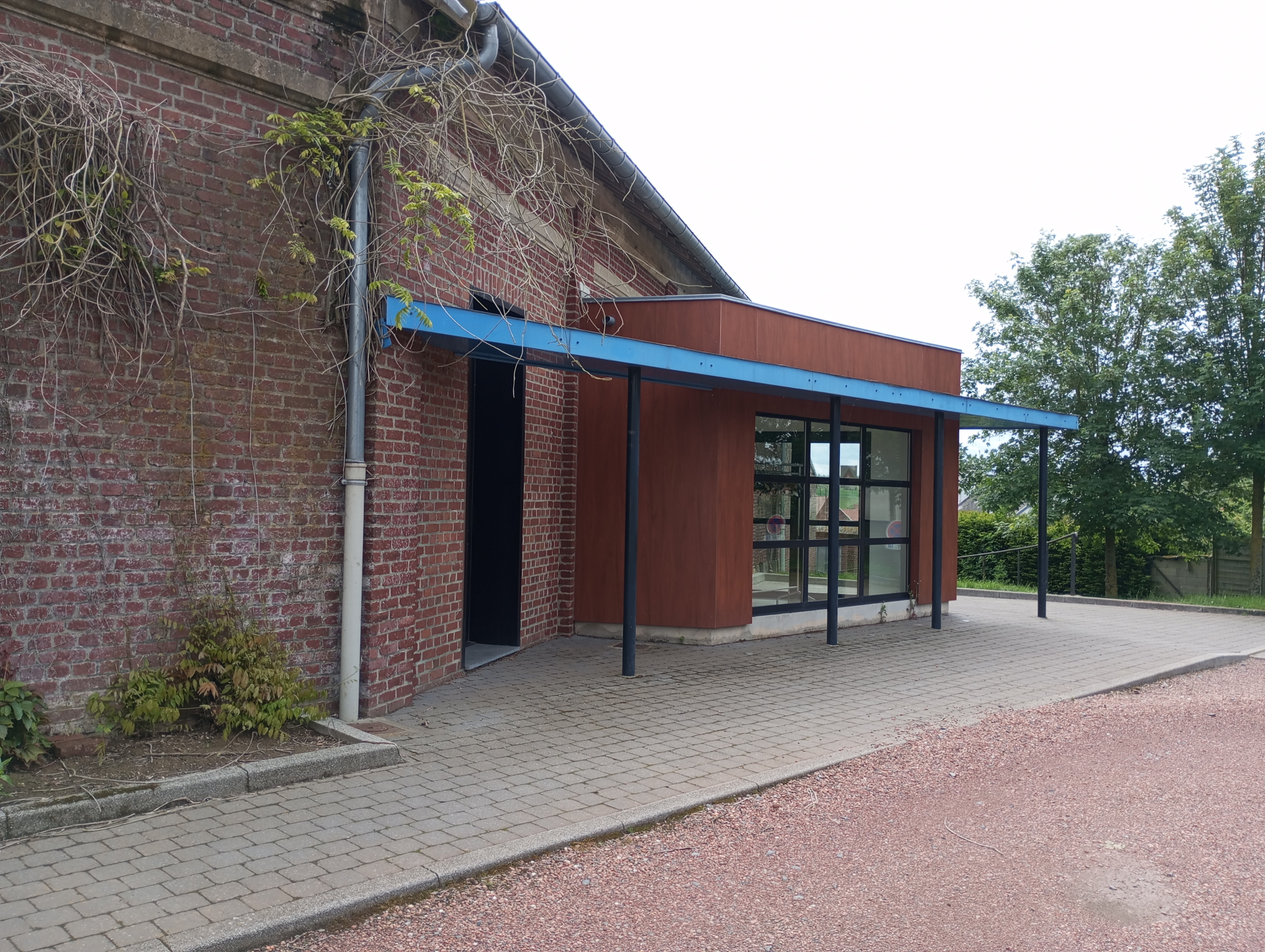 Bâtiment en brique avec entrée vitrée, auvent bleu, allée pavée et végétation.