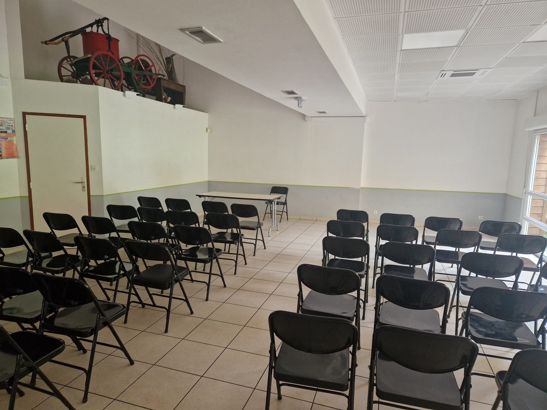 Salle avec rangées de chaises, table, porte, chariot rouge et blanc en hauteur, éclairage au plafond.