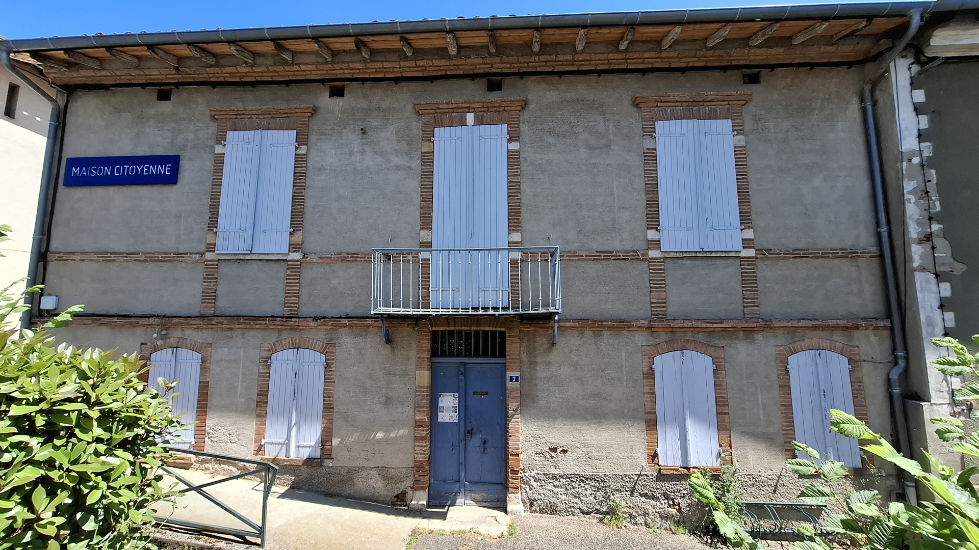 Bâtiment avec façade en briques, volets gris, balcon métallique, porte bleue et panneau "Maison Citoyenne".