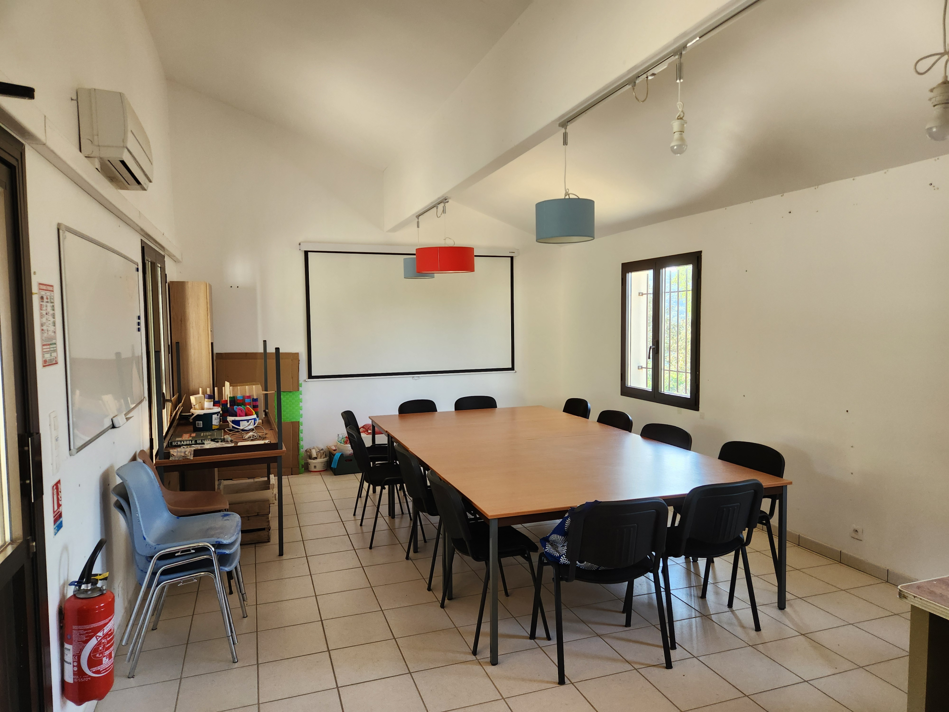 Salle avec table, chaises, tableau blanc, écran, ventilateur, et étagères.