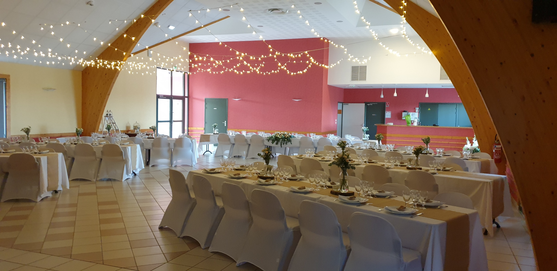 Salle avec tables, chaises, nappes, guirlandes lumineuses, vaisselle et fleurs.