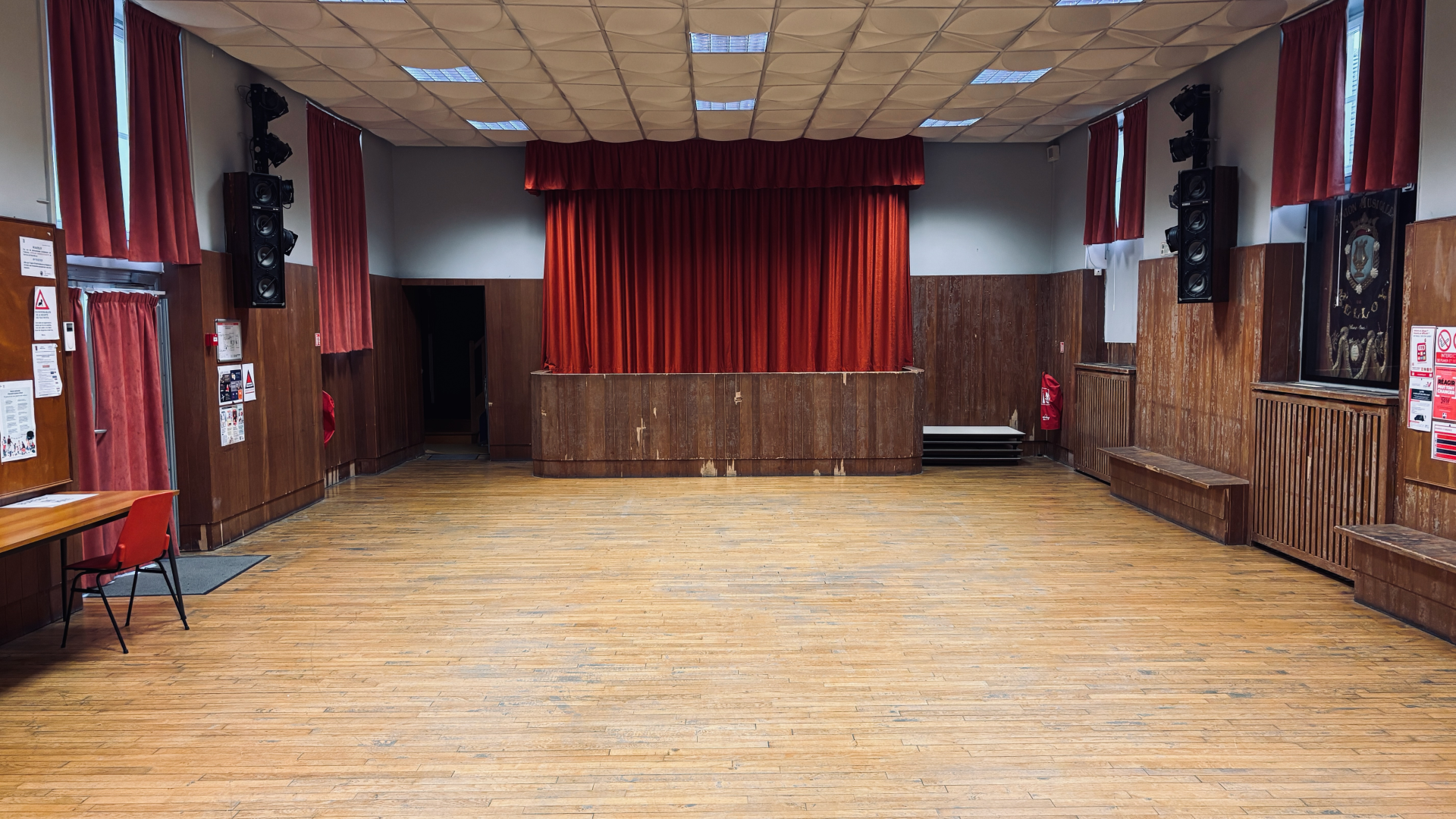 Salle avec scène, rideaux rouges, haut-parleurs, bancs et porte.