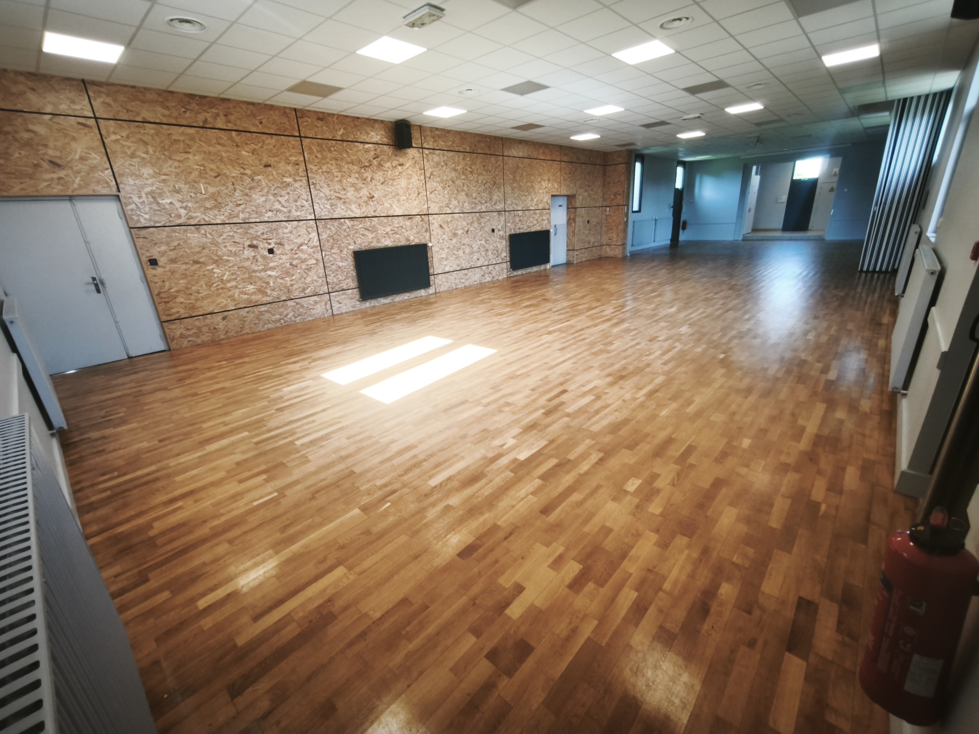 Salle vide avec parois en bois, sol en parquet, portes, extincteur et climatiseur.