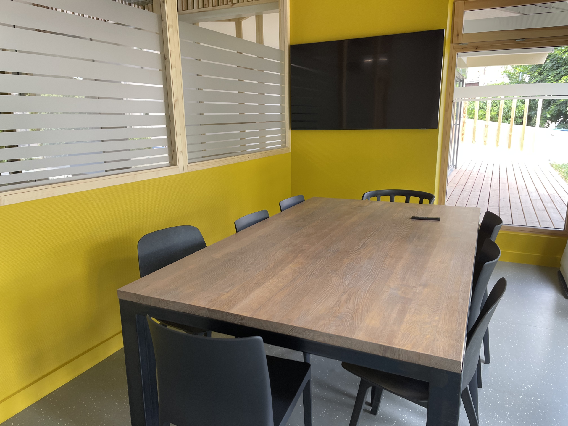 Table de réunion avec six chaises, mur jaune, fenêtres avec stores et porte ouverte.